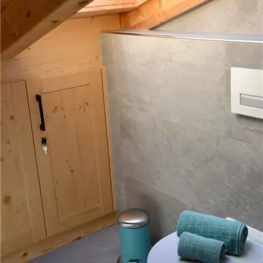 A modern bathroom in the attic with gray tiles and wood details. On the toilet are two turquoise towels and a small trash can is placed next to it.
