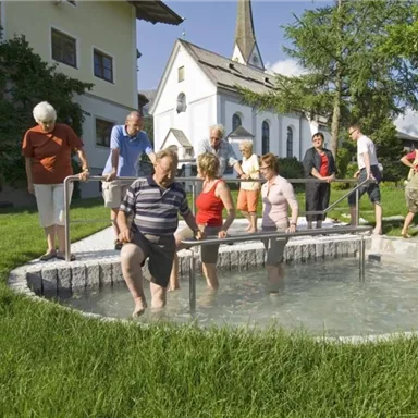 Eine Gruppe von Menschen steht um ein kleines Fußbad auf einer grünen Wiese. Im Hintergrund ist eine Kirche und blaue Himmel zu sehen.