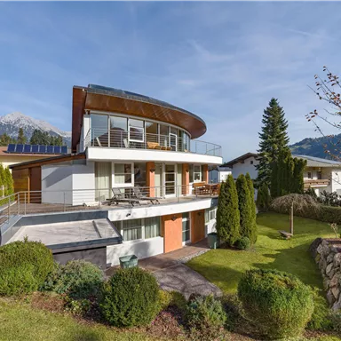 A modern house with a spacious terrace and large windows. The surroundings are green with trees and a view of the mountains.