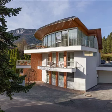 A modern house with large windows and a unique roof design. It is surrounded by trees and mountains nearby.