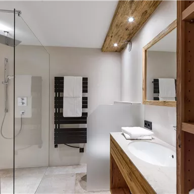 A modern bathroom with a glass shower and wooden accents. There are towels and a white sink area.