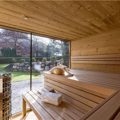 A cozy sauna room made of wood with a large glass wall overlooking a garden. Inside, there is a wooden bench and a towel ready.