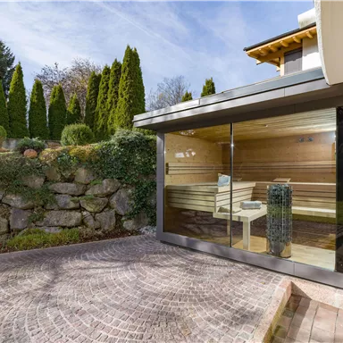 A modern sauna with large glass windows, surrounded by a well-maintained garden landscape. In the background, tall plants and a beautiful sky are visible.