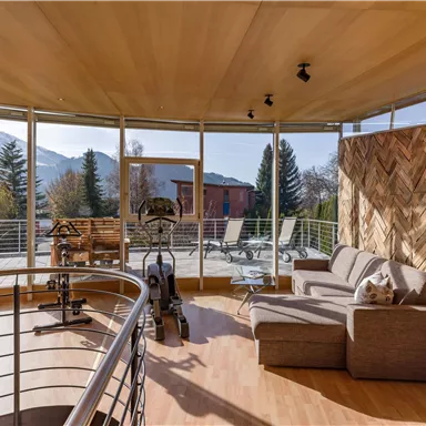A bright, modern living room with large windows and a view of the mountains. The room is furnished with a sofa, fitness equipment, and a wooden wall panel.