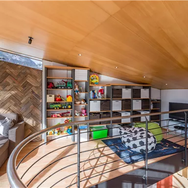 A modern attic room with a cozy sofa and an open shelf full of toys. The room has bright wood and plenty of daylight.