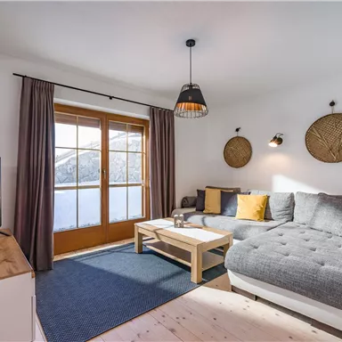 A cozy living room with a gray sofa and a wooden slatted table.  
Large windows provide a view of the surroundings.