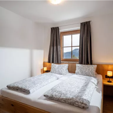A cozy bedroom with two beds and a large window front. The decor is modern and bright, with wooden details and soft colors.