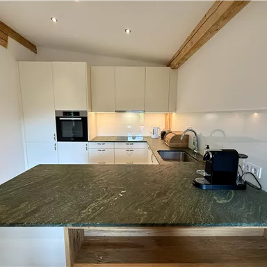 A modern kitchen with light cabinets and a large green workspace. The kitchen equipment includes a stove, a sink, and a coffee maker.