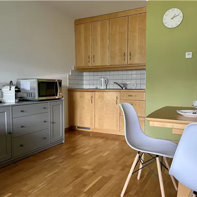 Eine helle Küchenecke mit Holzschränken und modernen Geräten. Ein Tisch mit Geschirr steht neben den Stühlen.