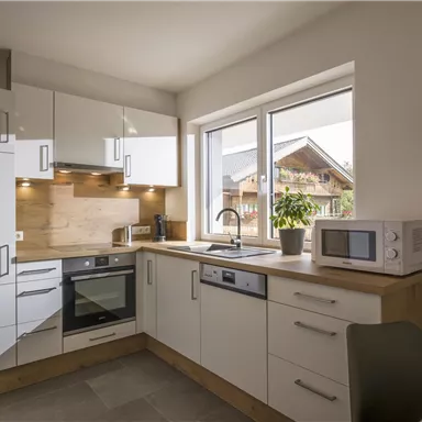 Eine moderne Küche mit weißen Schränken und Holzdetails. Am Fenster ist viel Tageslicht, und die Ausstattung umfasst einen Ofen und eine Mikrowelle.