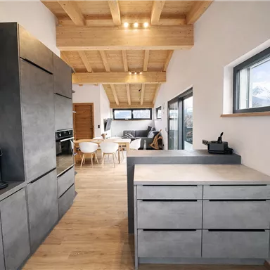 Eine moderne Küche mit betongrauen Möbeln und hellen Holzdecken. Im Hintergrund sind ein Essbereich und ein gemütliches Wohnzimmer sichtbar.