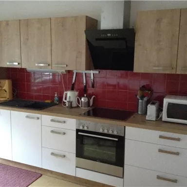Eine moderne Küche mit Holzschränken und roter Fliesenwand. Auf der Arbeitsfläche stehen Küchengeräte wie ein Wasserkocher und eine Mikrowelle.