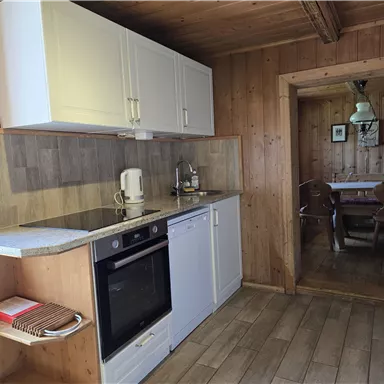 Eine moderne Küche mit Holzverkleidung, ausgestattet mit Herd, Spülmaschine und Wasserkocher. Im Hintergrund ist ein Essbereich sichtbar.