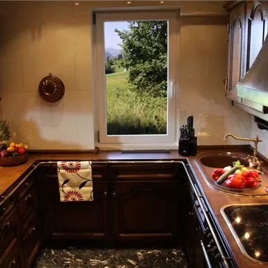 A modern kitchen with wooden cabinets and a large window front overlooking a green meadow landscape. Fresh vegetables and fruits, as well as a bottle of wine, are placed on the countertop.