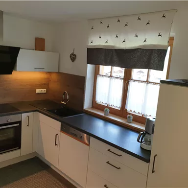 A modern kitchen with white cabinets and wooden details. It features a stainless steel sink and large windows that let in plenty of light.