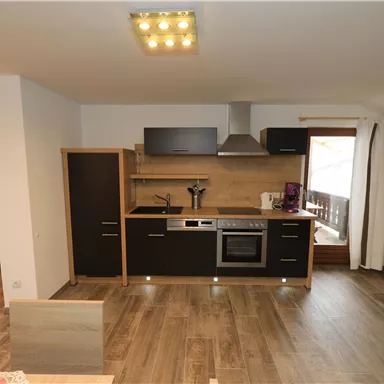 A modern kitchen with wooden flooring and elegant black cabinets. The window offers a view of a balcony.