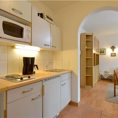 A small, modern kitchen with white cabinets and amenities like a microwave and kettle. In the background, a cozy living area is visible.