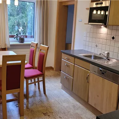 A modern kitchen with a bright decor and an adjoining dining table. The furniture is in warm wood and red tones.
