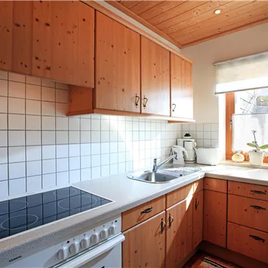 Eine helle Küche mit Holzschränken und einem modernen Herd. Das Fenster lässt viel Sonnenlicht herein und dekorative Gegenstände sind auf der Fensterbank platziert.