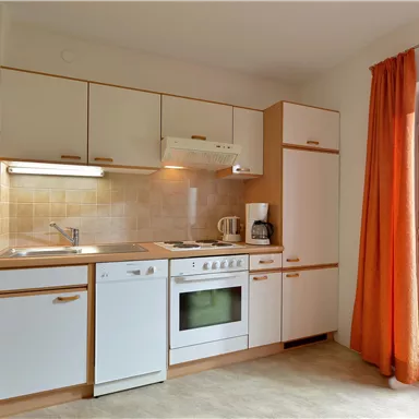 Modern kitchen with white cabinets and stainless steel appliances. A large window with orange curtains provides plenty of light.