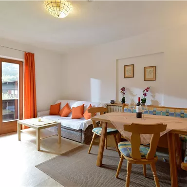 A cozy living space with orange curtains and a dining area. There is a sofa with cushions and a table in the center.