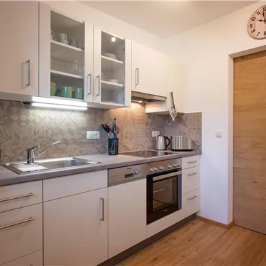 Eine moderne Küche mit hellen Schränken und einer Edelstahlspüle. Sie enthält einen Herd, einen Kühlschrank und eine Holztür.