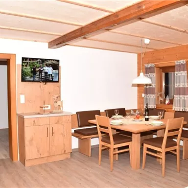 Ein gemütlicher Essbereich mit einem Holztisch und Stühlen. Im Hintergrund ist eine Küche und ein Fernseher sichtbar.