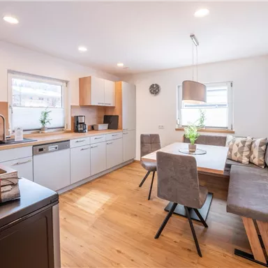 A modern kitchen with wooden surfaces and ample storage space. The dining area consists of a table and comfortable seating, surrounded by daylight.