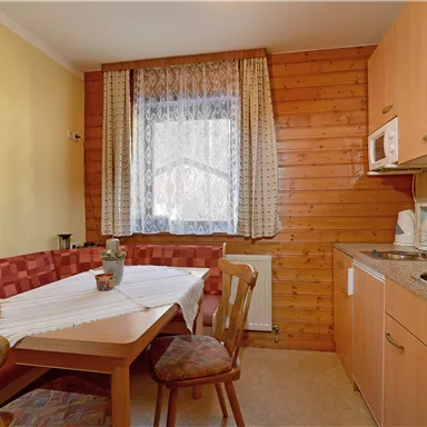 A cozy kitchen with a wooden table and chairs. The walls are made of wood and the room has large windows with curtains.