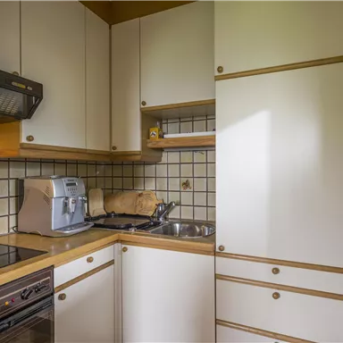 A small kitchen with modern appliances and light cabinets. The walls are covered with tiles, and there is a stove as well as a sink.