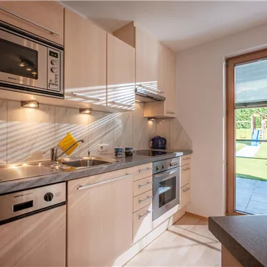 A modern kitchen with wooden cabinets and built-in appliances. Through the window, you can see a garden area with seating.