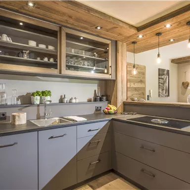 A modern kitchen with gray cabinets and wood details. There is a sink, a cooking area, and bright lighting.