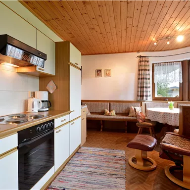 A cozy kitchen with a wooden ceiling and modern appliances. On the right side, there is a seating area with a window and a carpet.
