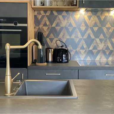 Eine moderne Küche mit einem eleganten Spülbecken und einem goldenen Wasserhahn. Im Hintergrund sind dekorative Fliesen in geometrischem Muster und verschiedene Küchengeräte zu sehen.