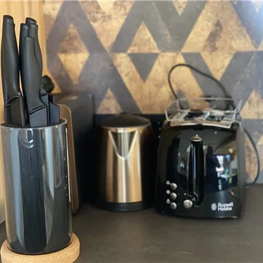Eine moderne Küchenecke mit einem Messerblock, einem Kaffeekocher und einem Toaster. Die Wände sind mit einem geometrischen Muster dekoriert.