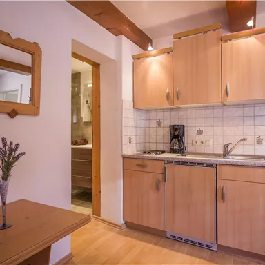 A cozy kitchen with wooden furniture and modern appliances. The walls are painted bright, and a small table with a vase is visible.