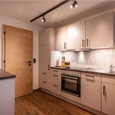 Eine moderne Küche mit hellen Schränken und einer Arbeitsfläche. Der Boden ist aus Holz und es gibt einen großflächigen Fensterbereich mit Vorhängen.