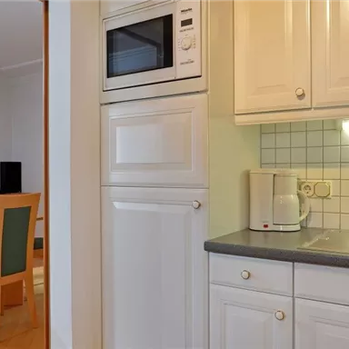 A small, modern kitchen with white cabinets and a sink. In the background, there is a dining area with a table and chairs.