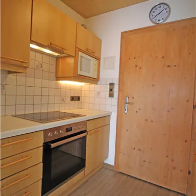A modern kitchen with wooden cabinets and a cooking area. The door is made of wood and a clock hangs on the wall.