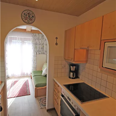 A modern kitchen with wooden cabinets and light tiles. In the background, there is an entrance to a cozy living area.