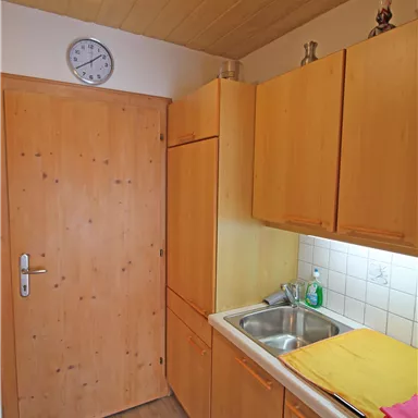 A small kitchen with wooden cabinets and a sink. A clock hangs on the wall and colorful towels are laid out on the countertop.