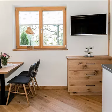 Ein modernes Wohnzimmer mit hellem Holzdekor und einem Esstisch. Der Raum verfügt über große Fenster und einen Fernseher an der Wand.