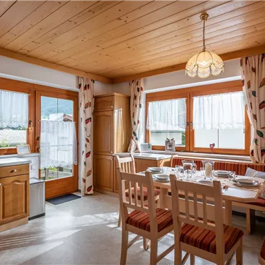 Ein gemütlicher Essbereich mit Holzdekor und großen Fenstern. Der Tisch ist für eine Mahlzeit gedeckt, und im Hintergrund sind Pflanzen und Dekoration zu sehen.