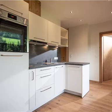 A modern kitchen with white cabinets and a built-in oven. The floor is made of wood and there are windows that provide plenty of light.
