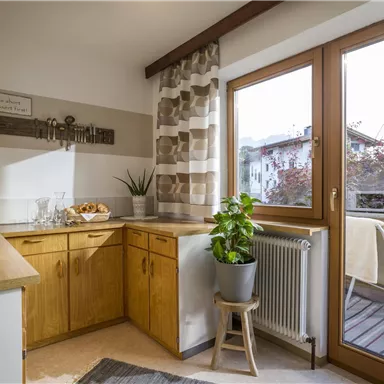 Eine helle Küche mit Holzschränken und großen Fenstern. Auf dem Tisch sind eine Pflanze und einige Snacks zu sehen, während ein Balkon im Hintergrund sichtbar ist.