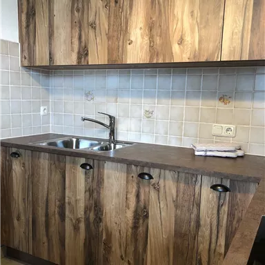 A modern kitchen with wooden fronts and a stainless steel sink. The floor is tiled and the walls are covered with white tiles.