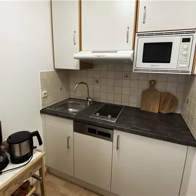 A small kitchen with a sink, an electric stove, and a microwave. Next to the kitchen counter are a coffee pot and a wooden board.