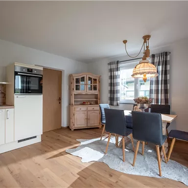 A modern kitchen with a dining table and chairs. The room design is bright and friendly, with wooden flooring and a short curtain.