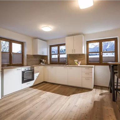A modern kitchen with white cabinets and large windows. The floor is made of wood and the room feels bright and inviting.