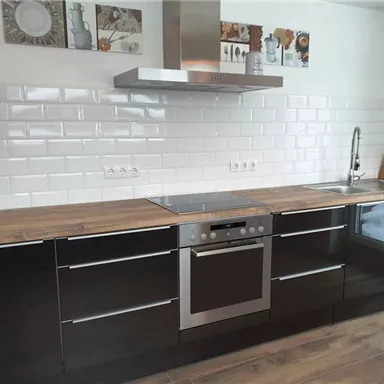 A modern kitchen with black fronts and a wooden countertop. White tiles are decoratively arranged on the wall along with pictures.